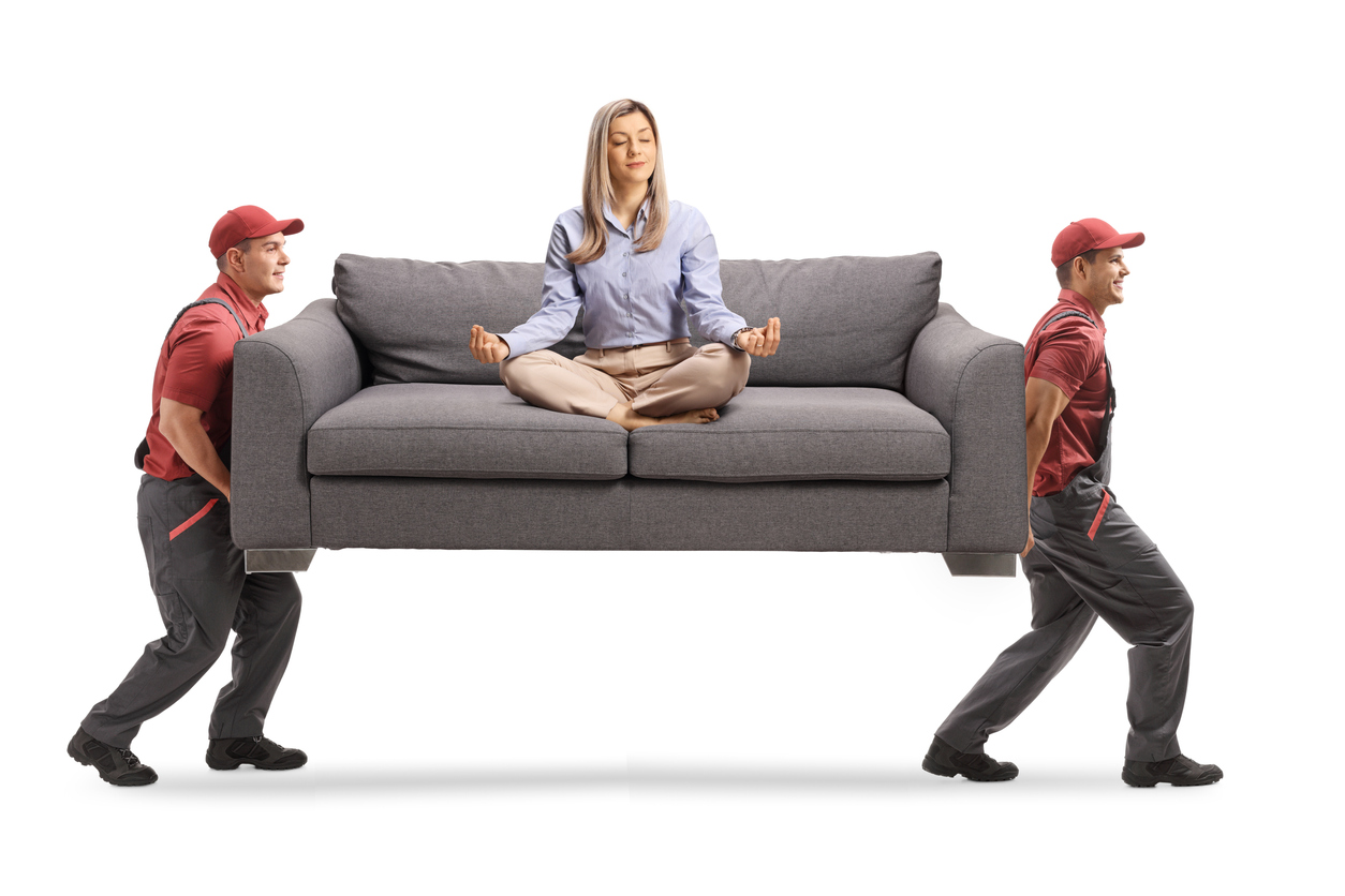 Young woman meditating on a sofa and two male workers carrying the sofa